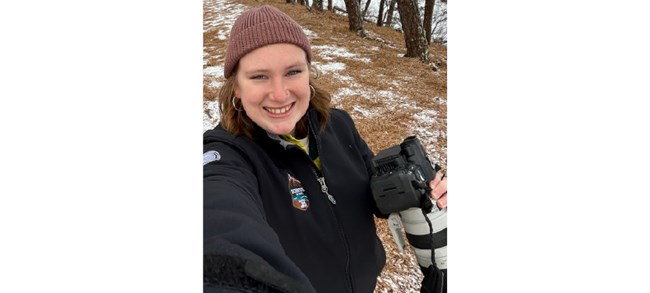 Perrault in a selfie as she holds a large camera-lens combo on a lightly snowy trail.