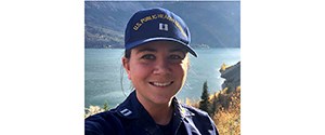 Headshot of Kayla in a navy and yellow cap, smiling in front of a mountain lake.