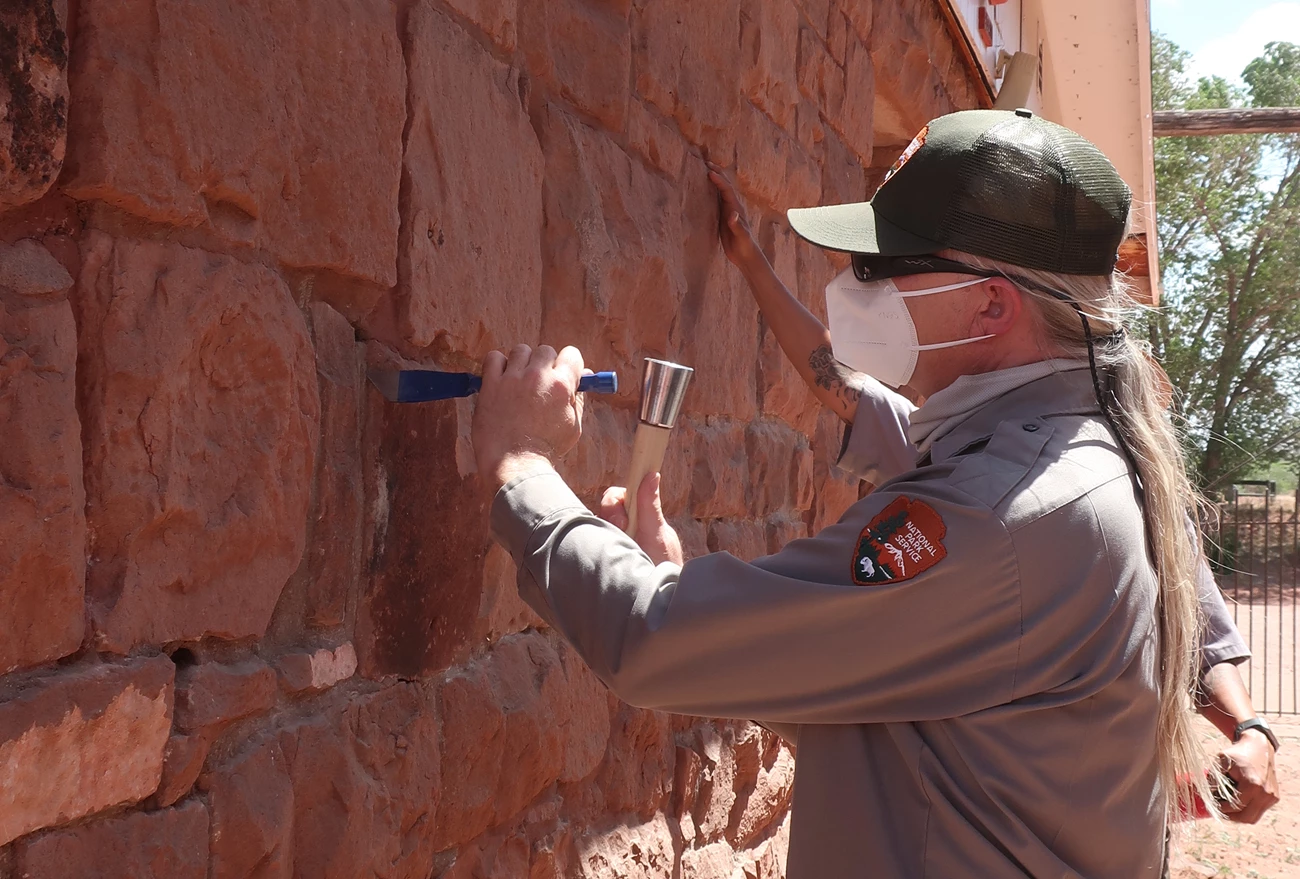 Keith Lyons taking a mortar sample at Hubble Trading Post. A man in National Park Service uniform, hat, and N95 mask uses a hammer and chisel to take a mortar sample from an old brick building.