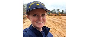 Headshot of Kelly outside in a navy and yellow cap, smiling at the camera.