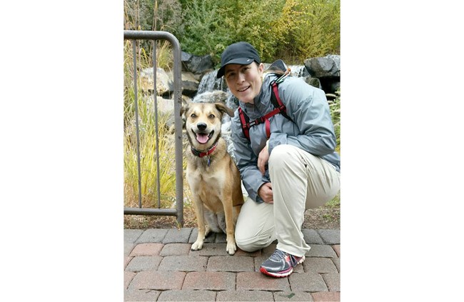 Woman in black baseball cap, blue jacket, and white pants kneels next to a tan-colored dog with a red collar.