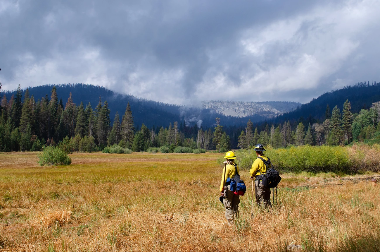 Burned Area Emergency Response Team in Lassen Volcanic after the Dixie Fire Two people in fire response uniforms look across a scenic field at smoke billowing from forested mountain foothills in the distance.