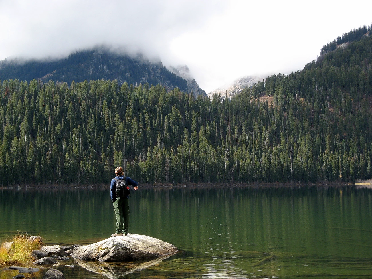 Lake in Grand Teton National Park A mountain lake surrounded by forest and steep mountain slopes. A man stands on a rock overlooking the lake.