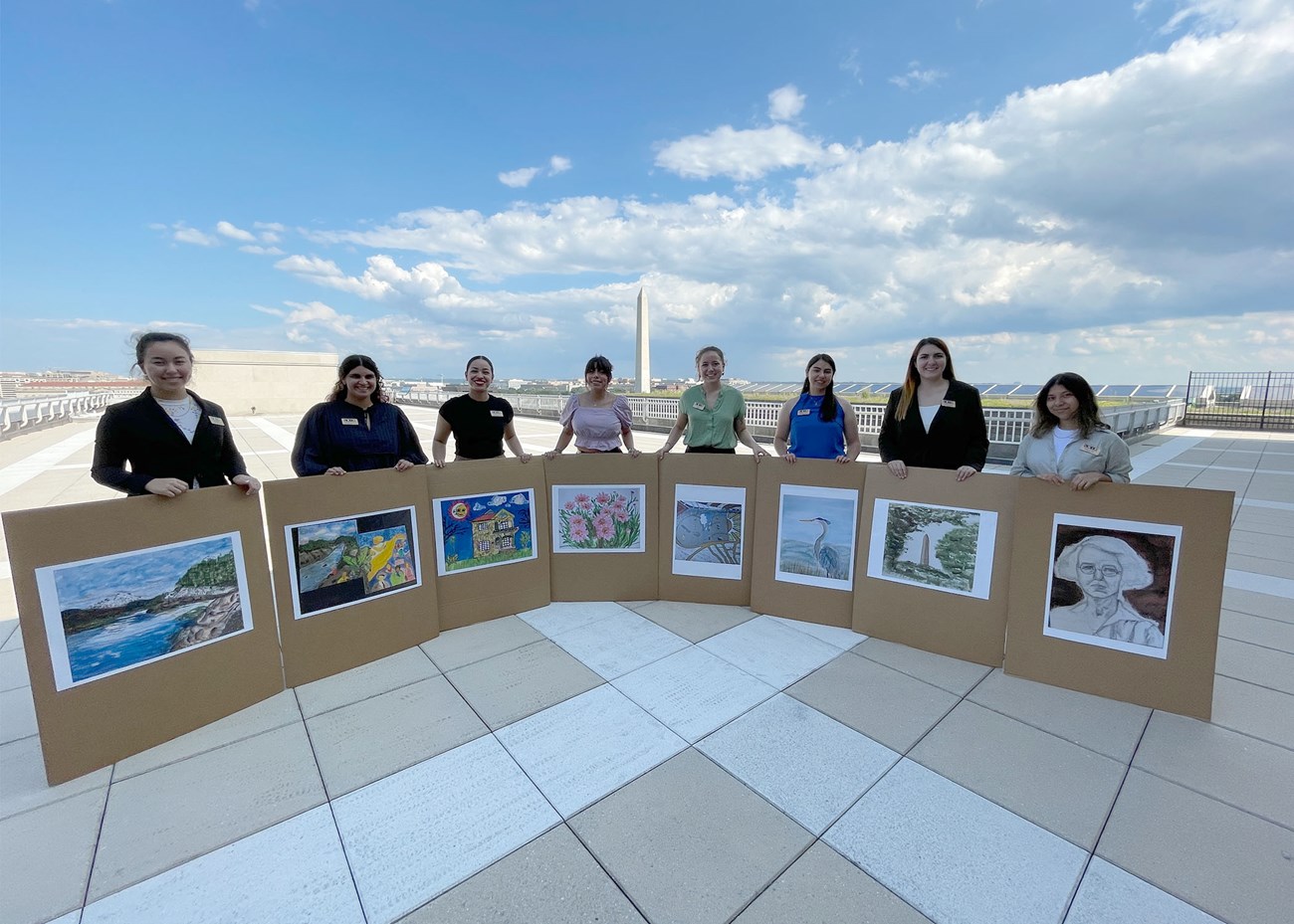 nine students holding pictures of their drawings and paintings with the washington monument in the background