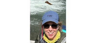 Lynne wearing a ball cap, smiling at the camera. There's a bear in a river behind her.