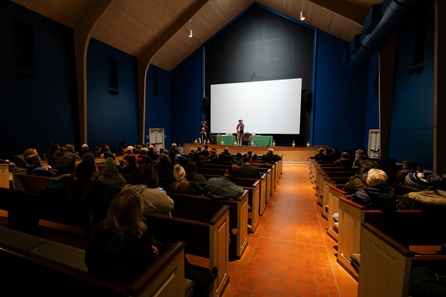 Presenters on a stage address an audience seated in an auditorium