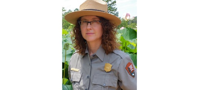 Milton outdoors in a National Park Service flat hat and gray shirt. She stands in front of towering wetland plants as tall as she is.
