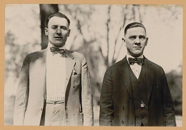 Two smiling men in suits and hats stand beside each other.