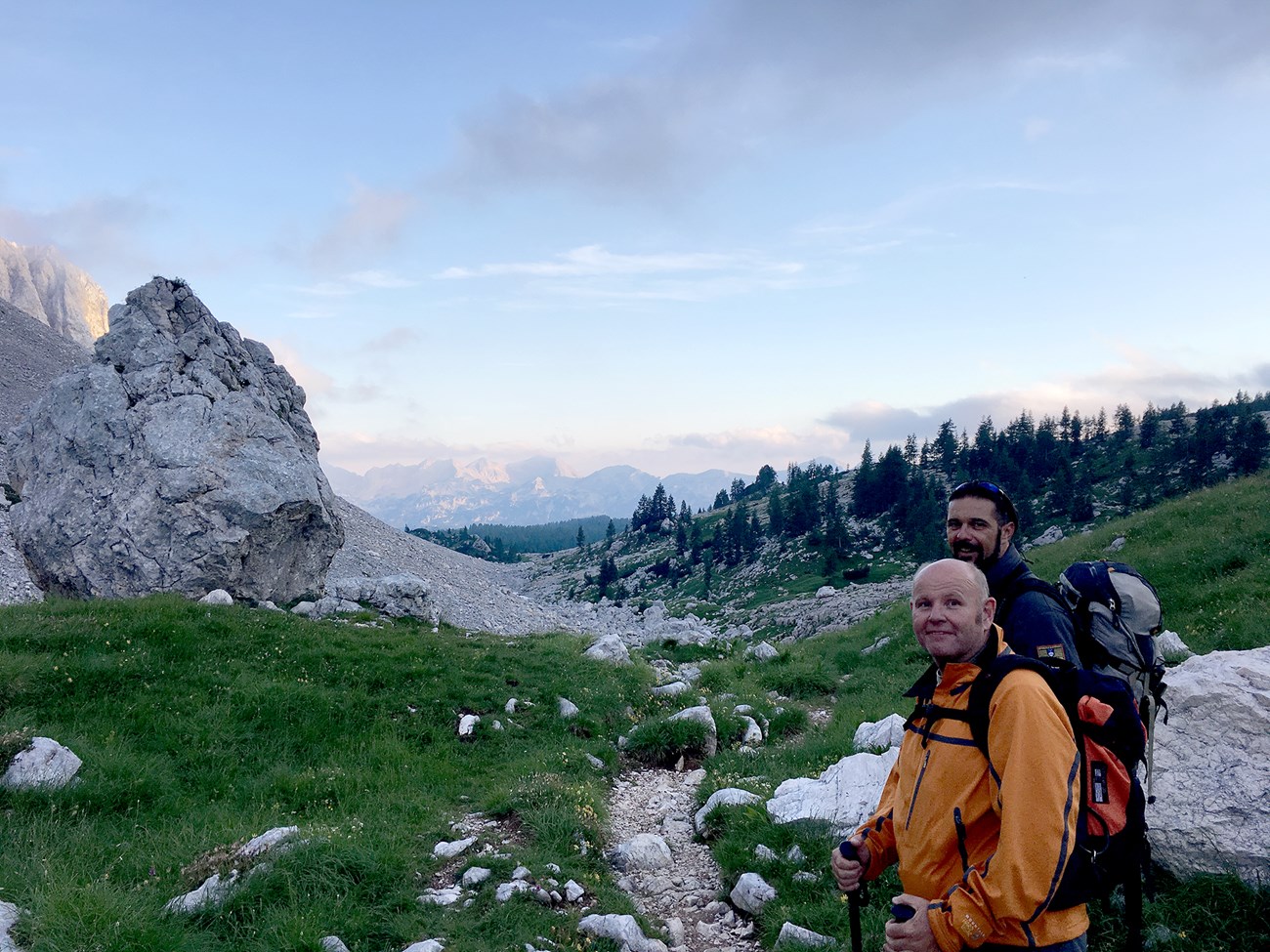 Two men stand in front of a green meadow with stony gray cliffs, evergreen trees, and distant pink and blue mountains