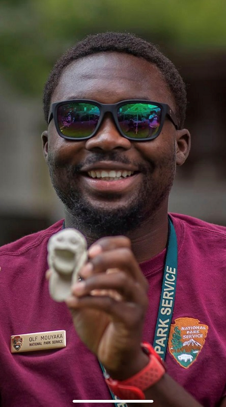 Olf Mouyaka Olf smiling to the camera holding a small sculpture of a person with an open mouth made out of rock