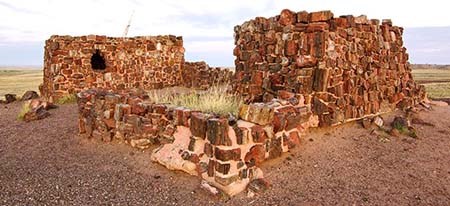 Agate House pueblo Two piles of rocks in reds and whites similar but larger than brick, arranged loosely into two dwellings, one with a window. A short perimeter of the same rocks appearing like a fence around the dwellings. Tall green grass grows inside he fence.