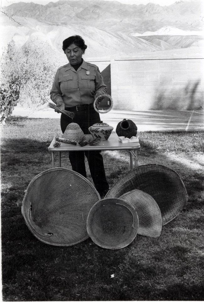 Native woman in a NPS park ranger uniform giving a talk. She is standing in front of a table with several native baskets.