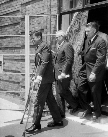 A black and white photo of President Kennedy in a suit, smiling as he walks on crutches out of a building, followed by three men in suits.