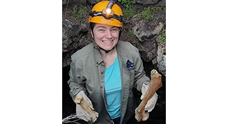 Rapp wearing a helmet and gloves as she stands up in a hole--a faunal trap--in a volcanic landscape holding up two large bones.