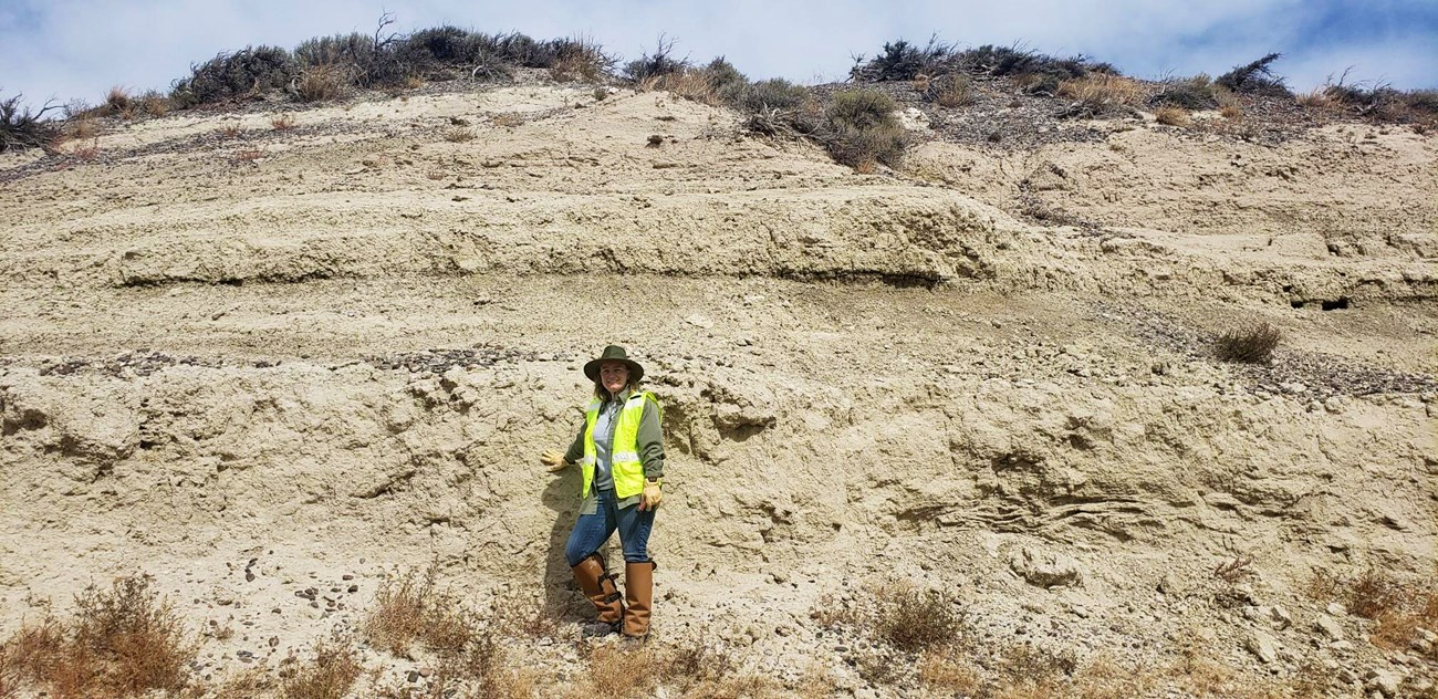 Woman wearing a wide brimmed hat, gloves, a yellow safety vest, and gaiters leaning against a bluff consisting of layers of lose, light-colored rock.