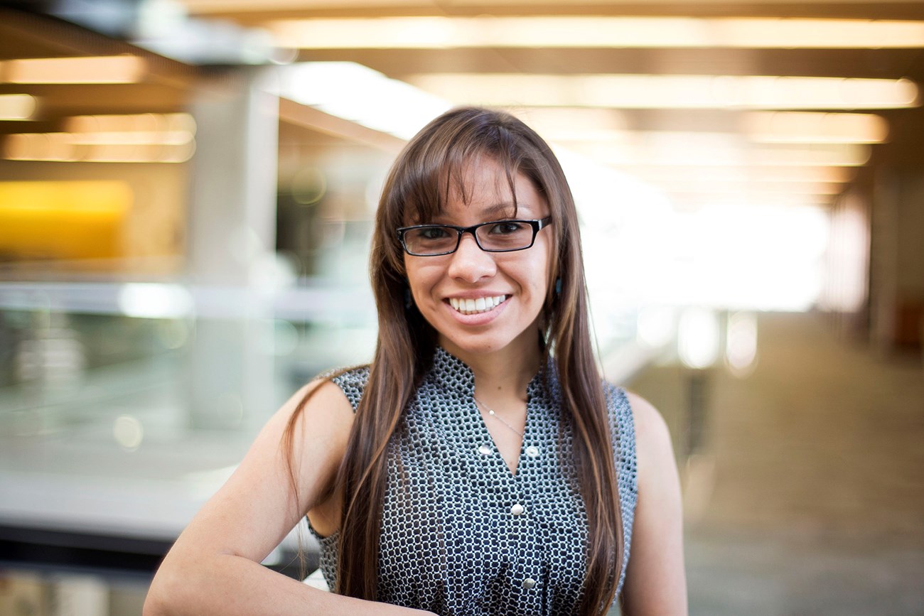 A young woman with long, dark hair and glasses, wearing a blue checkered top, smiles at the camera.