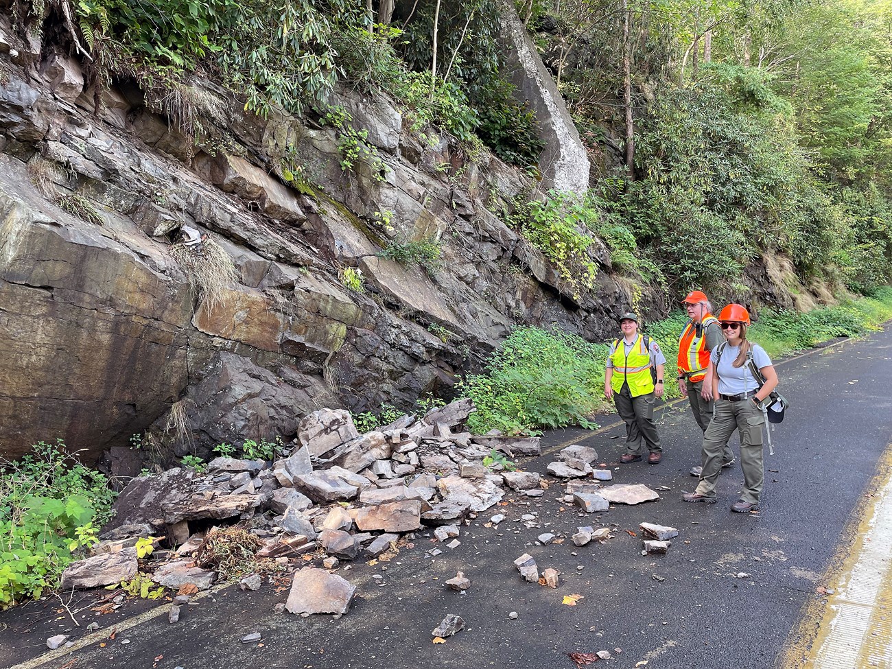 Three people in hard hats and safety vests stand on a road beside a rock fall, where part of a rocky cliff backing the road has crumbled onto it.