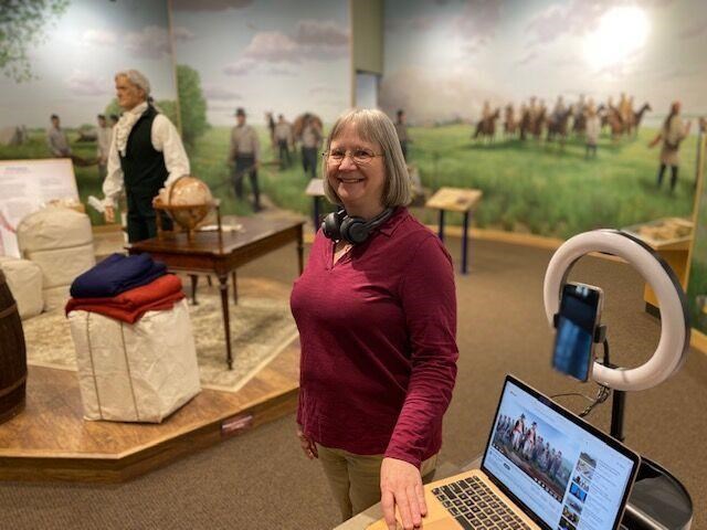 A woman stands with an open laptop and a ring light in a museum. The museum includes a mannequin wearing historic clothing and walls with murals of people with horses.