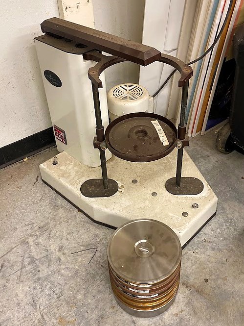 Big device with a round metal sieve tray-holder attached to two vertical metal bars connected at the top by a metal arm that can move both bars--and the tray--back and forth.