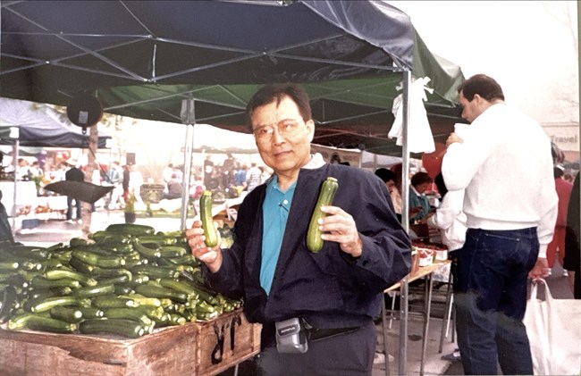 TS farmers market A man standing next to a produce stand holding two zuchinni.
