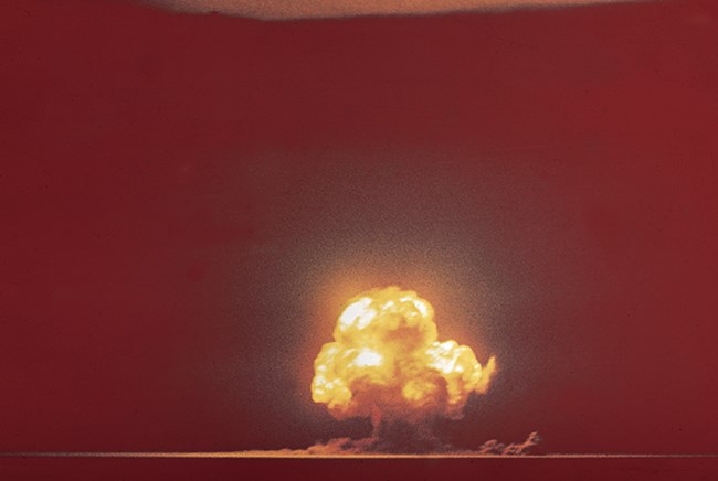 Color photograph of bright yellow mushroom cloud with smoke and dirt spewing at the horizon line. Background is a deep, dark red