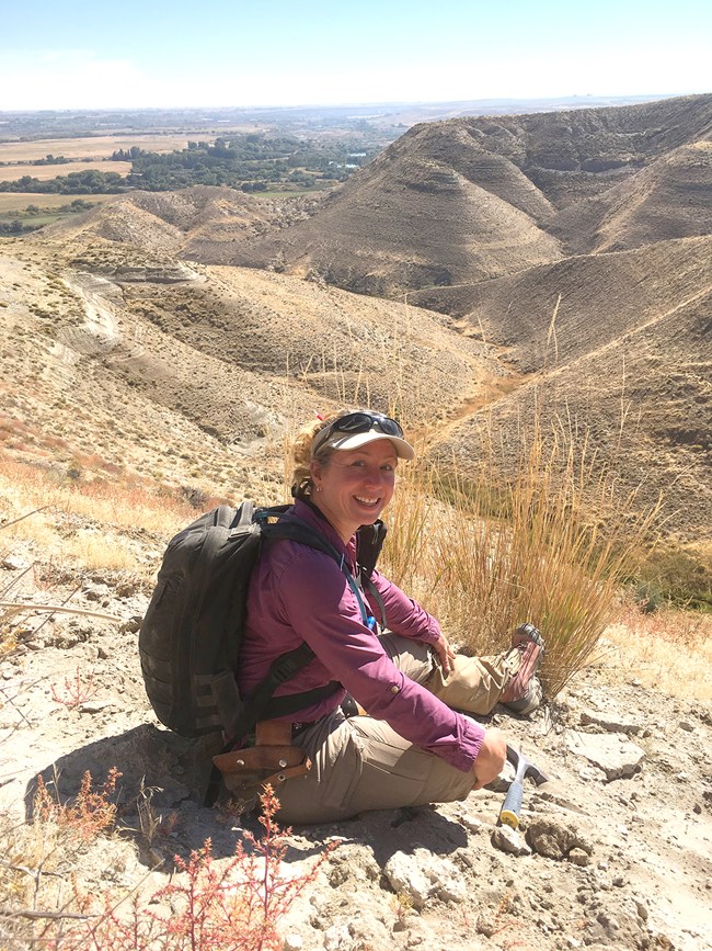 Woman with sun visor sits on hill overlooking bluffs. She is wearing a backpack, and a hammer lies in front of her.