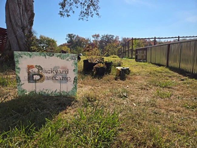 Outdoor area with a sign, tree stumps, and a greenhouse. Transcribed Text: Backyard Basecamp Outdoor area with a sign, tree stumps, and a greenhouse. Transcribed Text: Backyard Basecamp
