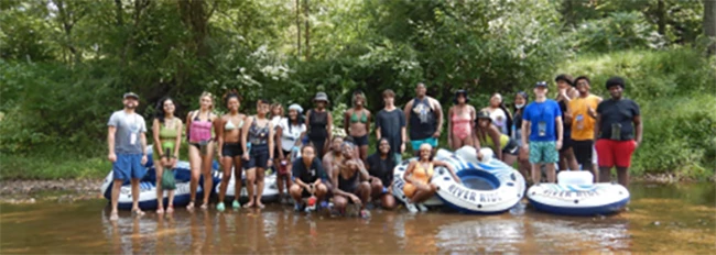 A group of 24 young people standing along the side of a creek with air filled innertubes for floating. There are trees in the background. A group of 24 young people standing along the side of a creek with air filled innertubes for floating. There are trees in the background.