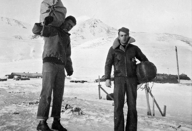 in a historic scene, two men stand dressed in jeans and leather jackets in the foothills of a mountain entirely covered in snow.