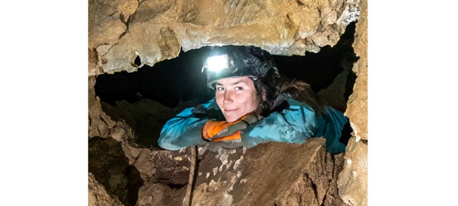 Heimel wearing gloves and a headlamp, lying on her stomach, resting her chin on her hands, and smiling down through a cave opening.