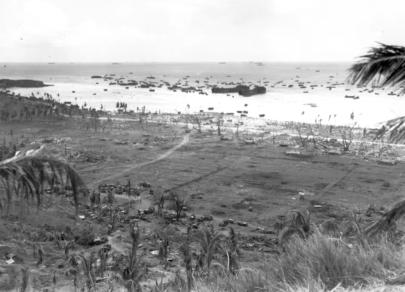 While it's best known as the northern beachhead during the Battle of Guam in 1944, Asan Beach is a site of rich cultural and historical significance. View overlooking a beach covered with twisted and burnt trees. At the edge of the beach are many trucks. The ocean is full of ships.