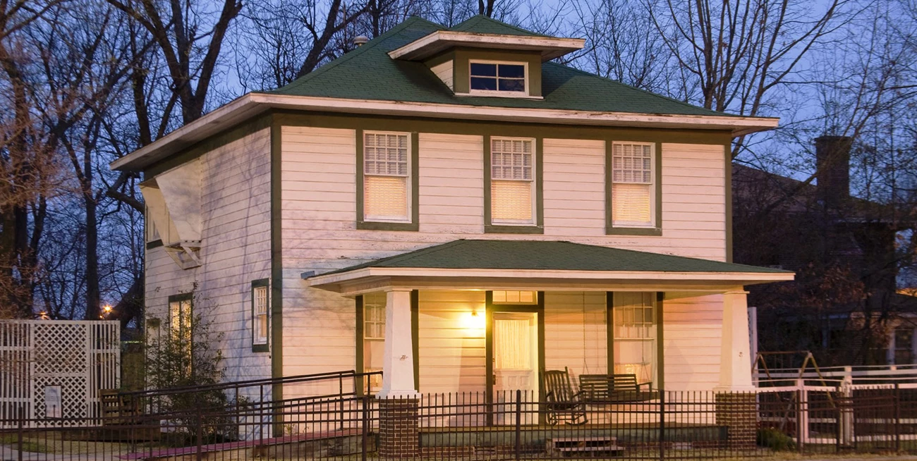 Birthplace home of William Jefferson Clinton White two story house lit at dusk with green trim and trees behind.