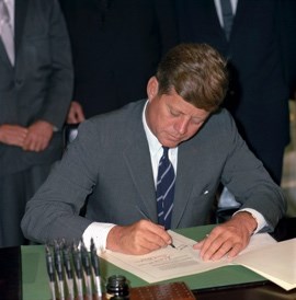 A photo of President Kennedy in a grey suit and blue tie sitting at a desk, signing a stapled group of papers on a green writing pad with a pen. Others in suits look on in the background. The pen stand, inkwell, and extra pens are in front.