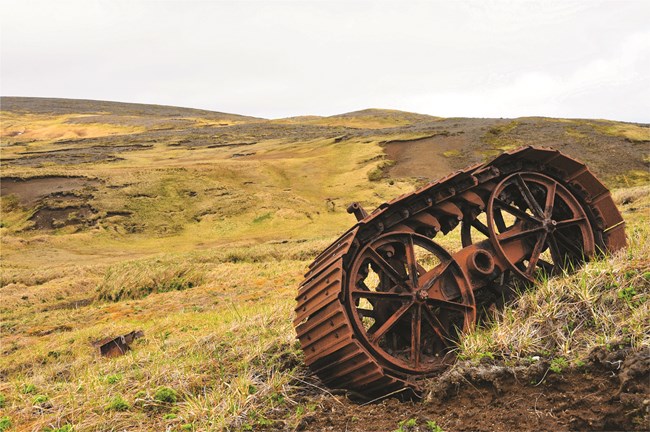 Color photo of grassy field, fog, and a rusted track assembly.