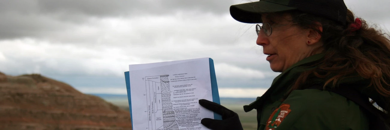 p a women with a ponytail and NPS baseball hat points at a geologic diagram while standing in front of badlands formations.