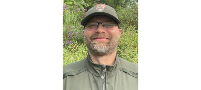Tzilkowski smiling outside in front of lush greenery in an NPS field uniform and glasses.