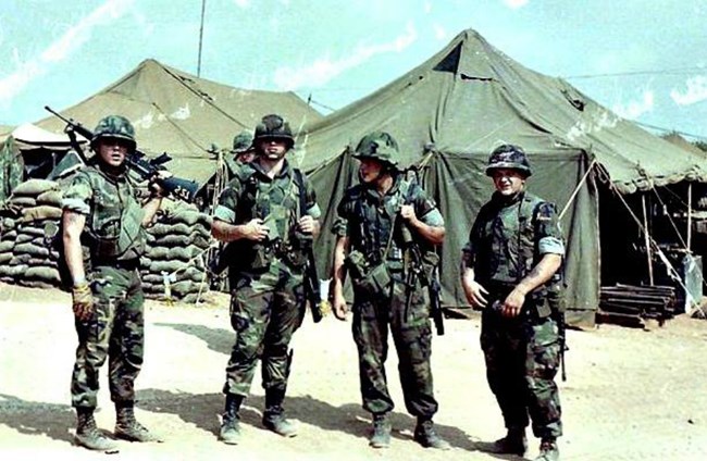 Four US Marines in combat fatigues stand in front of a wall of sand bags in a desert-like location.