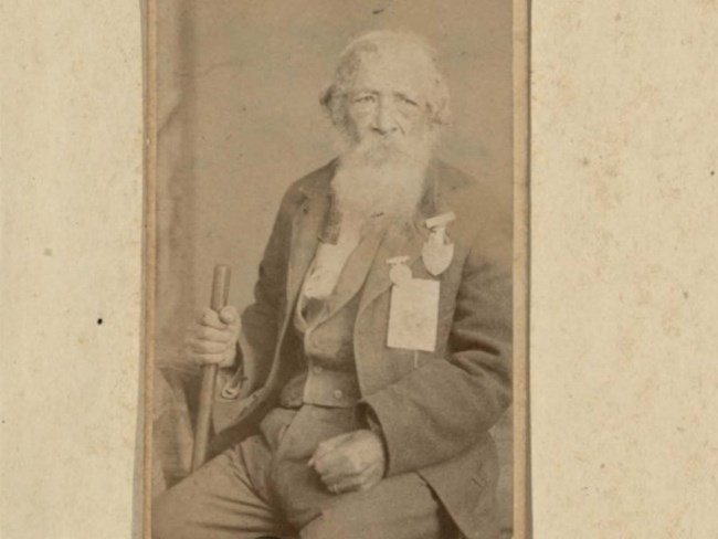 Jordan Noble, at around 87 years of age, sits for a full portrait in civil clothes, two medals of honor pinned to chest