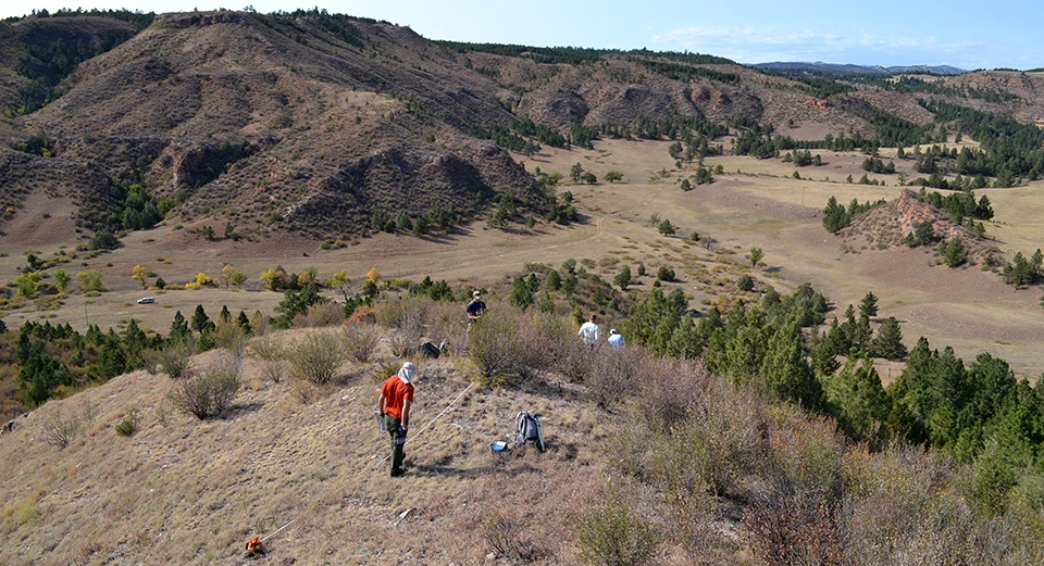 Plant community monitoring Two people standing by a measuring tape on a hilltop overlooking a valley and more hills, and two people walking down the hill.
