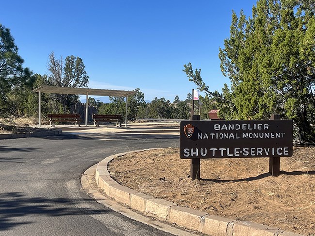 An image of a sign reading "Bandelier National Monument Shuttle Service" in a parking lot.