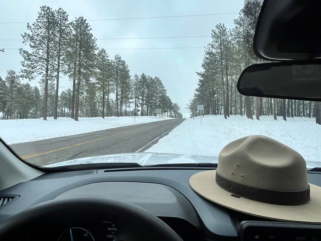 A ranger hat sits on the dashboard of a car. Looking through the windshield, the road is wet and snowy.