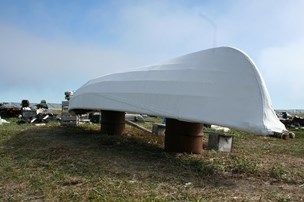 A modern white umiak boat sits face-down on two rusty barrels under a blue sky