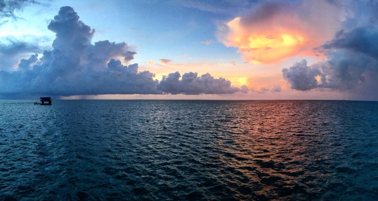 Brilliant orange sunset behind clouds. A small silhouette of a house on stilts rising above the bay waters in the distance.