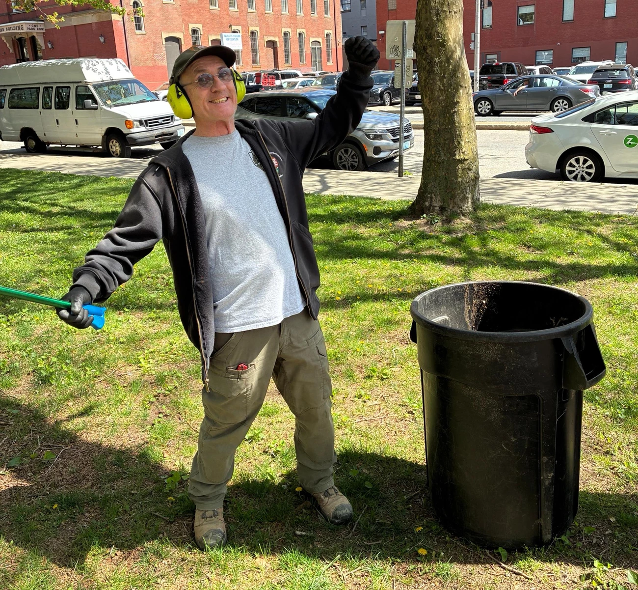Jay Man in black sweatshirt and wearing hat stands next to trash bucket with hands raised and a picker in one hand