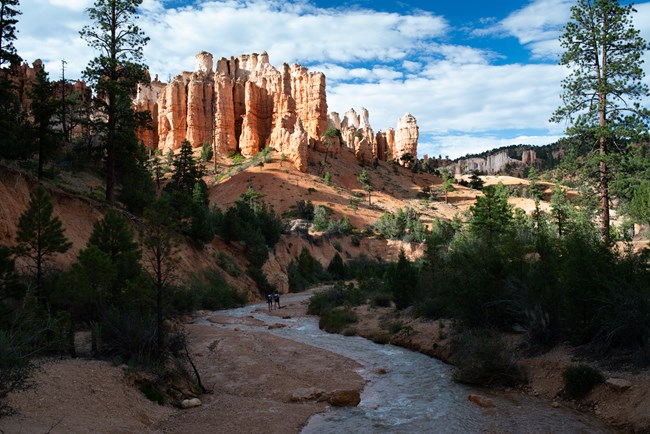 Two hikers walk along a river under a large rock formation