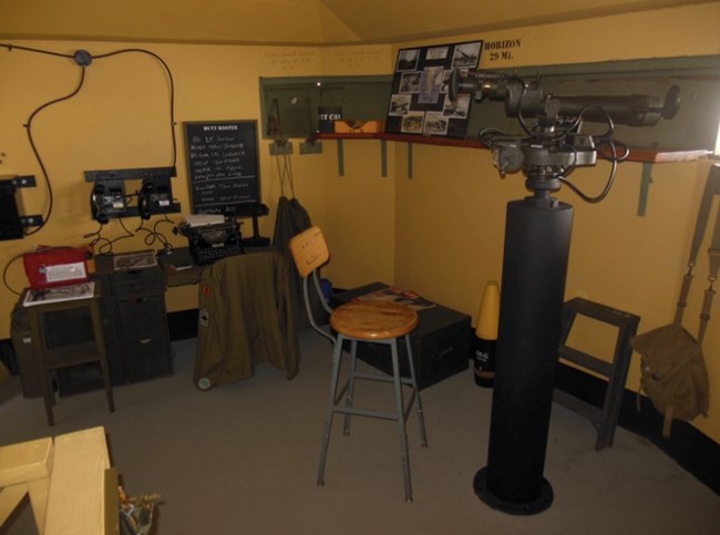 An observation room inside a WWII bunker with a large telescope on a pedestal, communication radios, a typewriter, and a chalkboard. A jacket is draped over a stool, and a shelf displays images and notes.