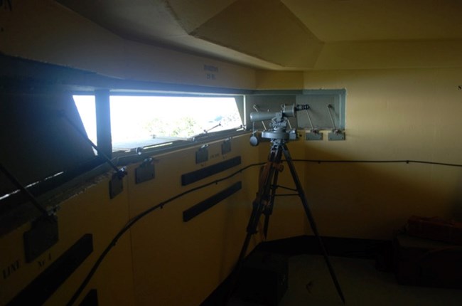 An interior view of a WWII observation station with a narrow observation window, a mounted instrument on a tripod, and labels on the wall. The setup provided an expansive view of the horizon for tracking enemy vessels.