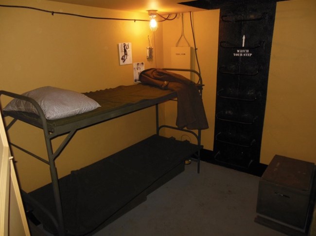 A bunkroom with two metal-framed bunk beds and a ladder on the wall labeled "Watch Your Step." A single light bulb hangs overhead, illuminating personal effects such as a pillow and a folded jacket on the top bunk.