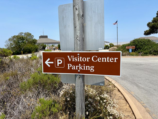Brown sign says "Visitor Center Parking" with a left arrow and parking symbol, near shrubs.