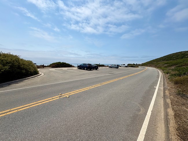 Curving road by a small parking lot, a few cars, ocean in the distance, blue sky, green hills.